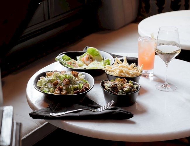 A table set with salad, fries, and drinks, including a glass of wine and a pink cocktail, inviting a cozy dining experience.