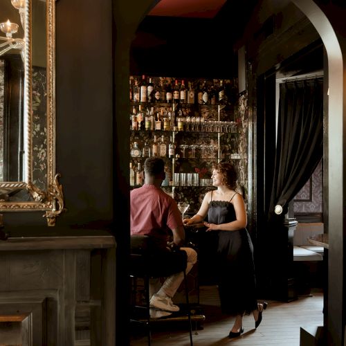 Two people sitting at a bar with a vintage style interior, including a mirror and fireplace, in a dimly lit room.