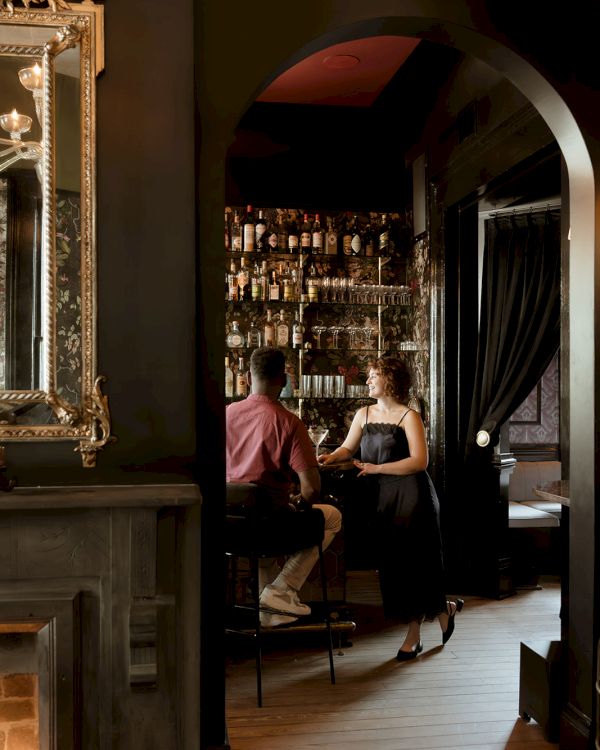 Two people sitting at a bar with a vintage style interior, including a mirror and fireplace, in a dimly lit room.