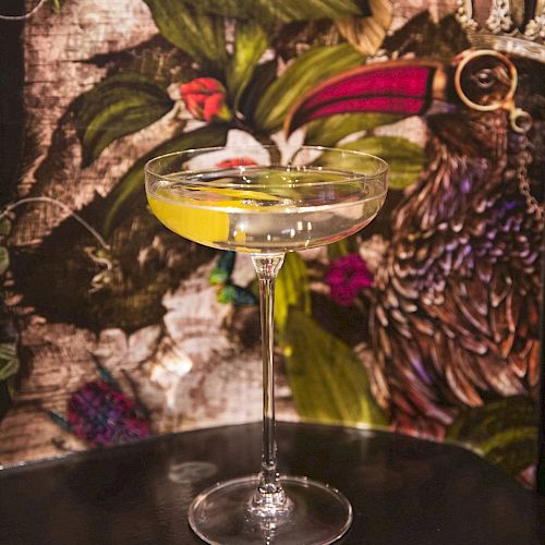 A cocktail in a coupe glass with a lemon twist, set against a floral and bird-themed background.