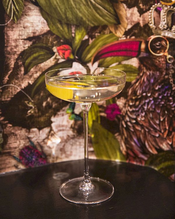 A cocktail in a coupe glass with a lemon twist, set against a floral and bird-themed background.