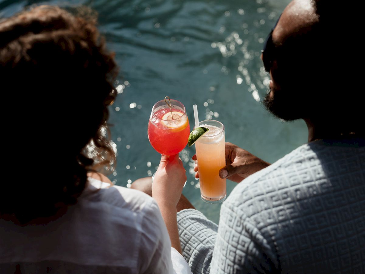 Two people relaxing by the water, holding colorful drinks with garnishes in their hands, creating a serene and refreshing atmosphere.