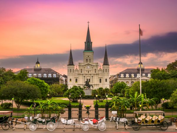 The image shows a sunset view of a historic building with spires, surrounded by greenery and horse-drawn carriages in front.