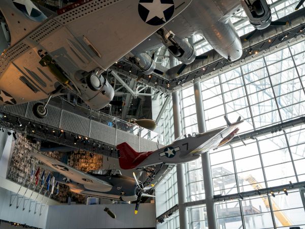 The image shows vintage military aircraft suspended from a museum ceiling with large windows and a display of flags in the background.