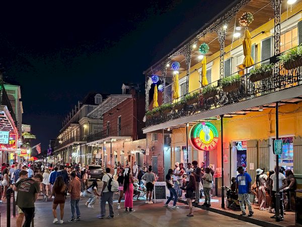 The image shows a bustling street scene at night, with people walking past vibrant buildings, neon signs, and decor, creating a lively atmosphere.