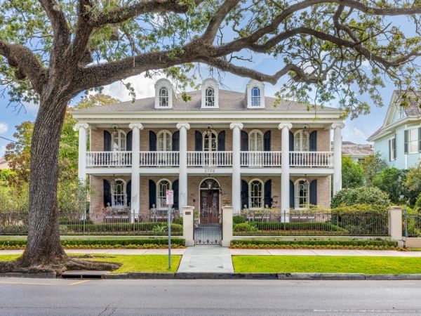 The image shows a large, elegant two-story house with white columns, symmetrical windows, and a manicured lawn, framed by a large tree.