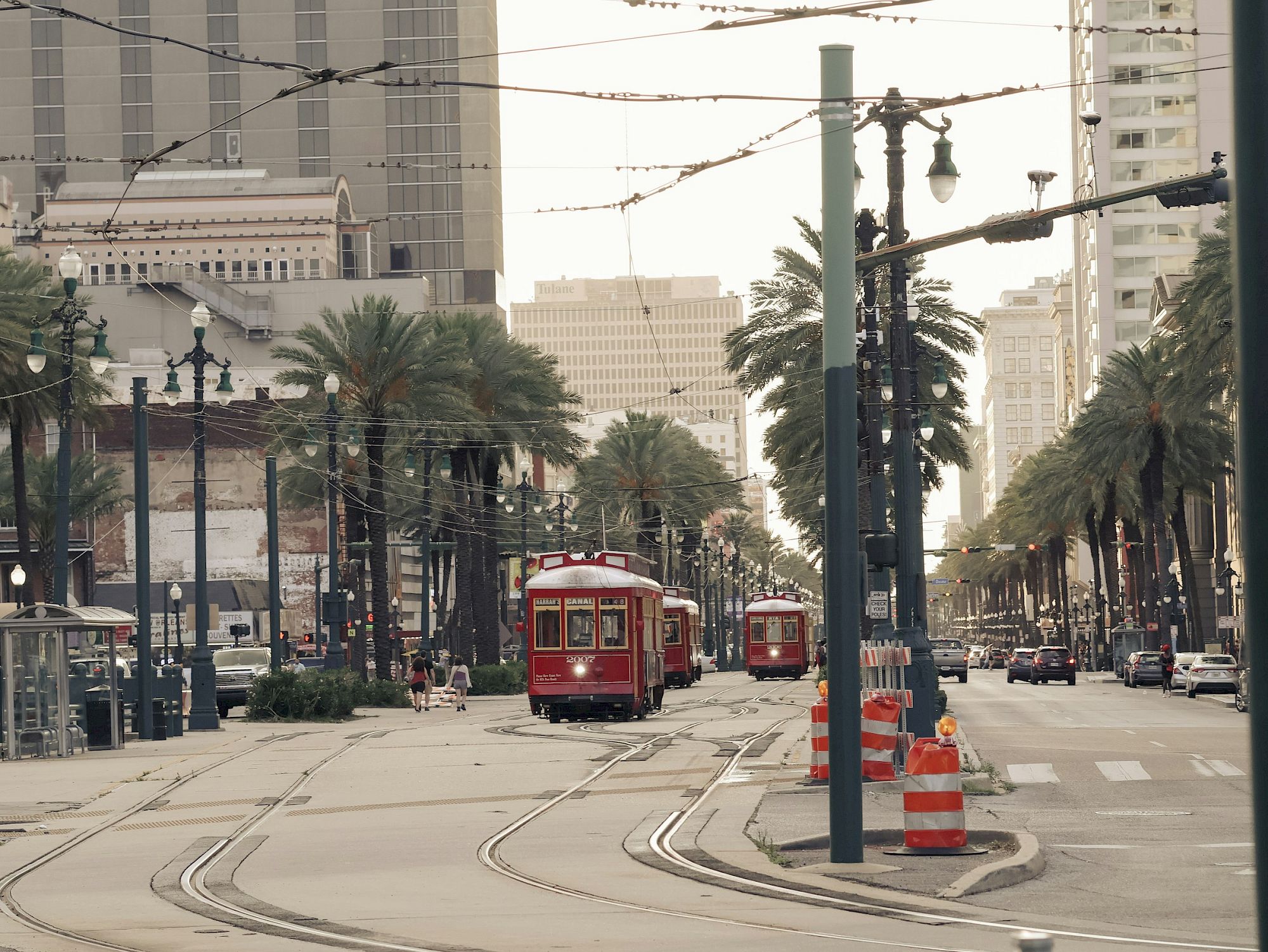 The image shows a city street with red streetcars, palm trees, tall buildings, and orange construction cones lining the road.