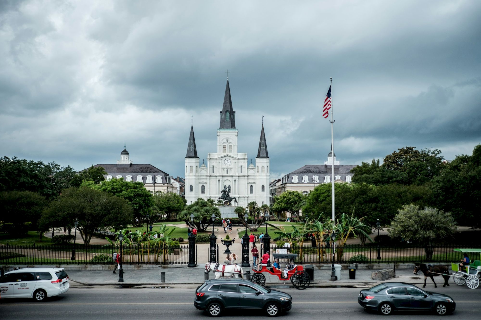 The image shows St. Louis Cathedral in New Orleans, with people, cars, and a horse-drawn carriage in front, under a cloudy sky.
