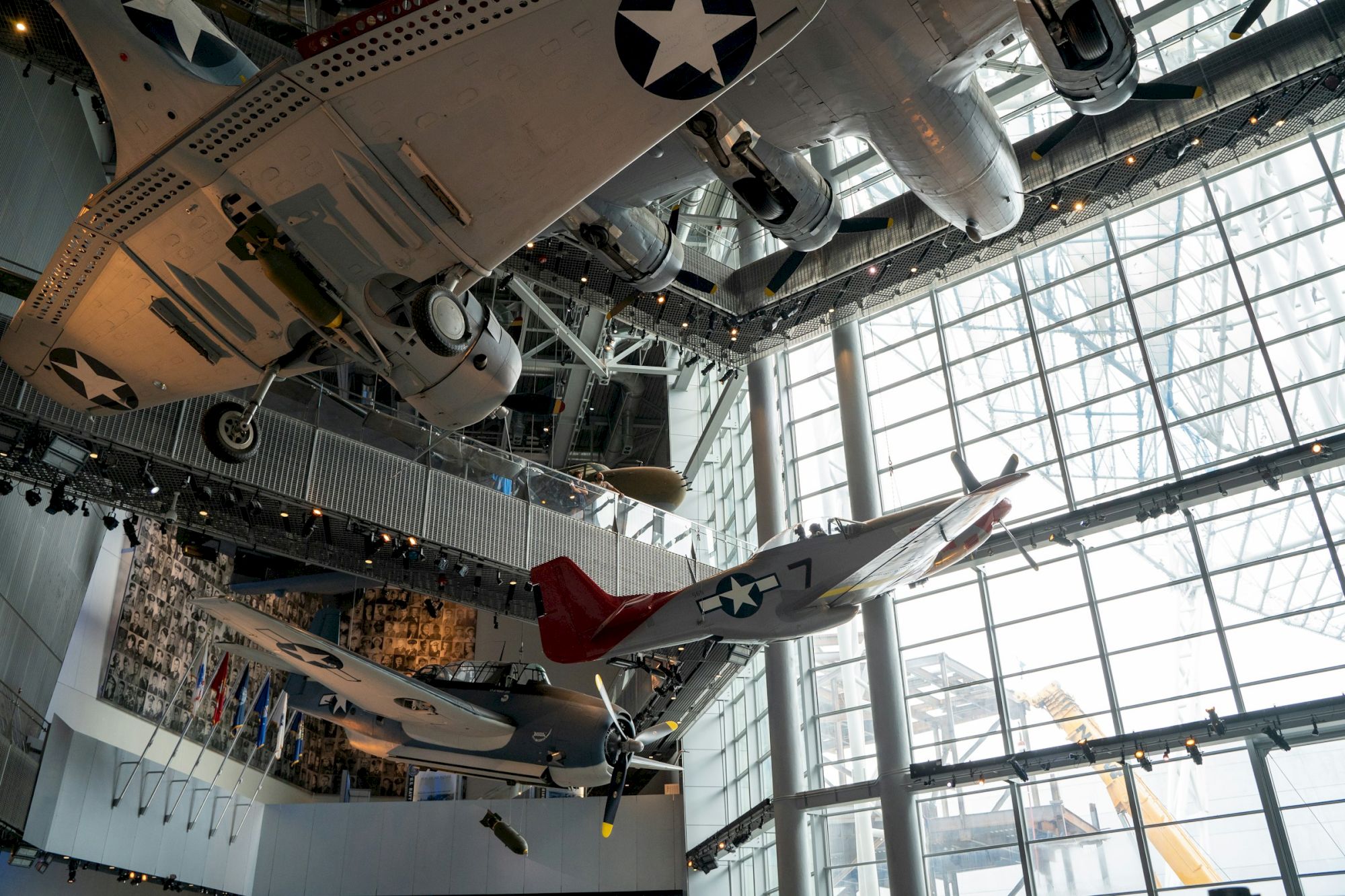 The image shows vintage military aircraft displayed indoors, suspended in a large glass-walled exhibit space.