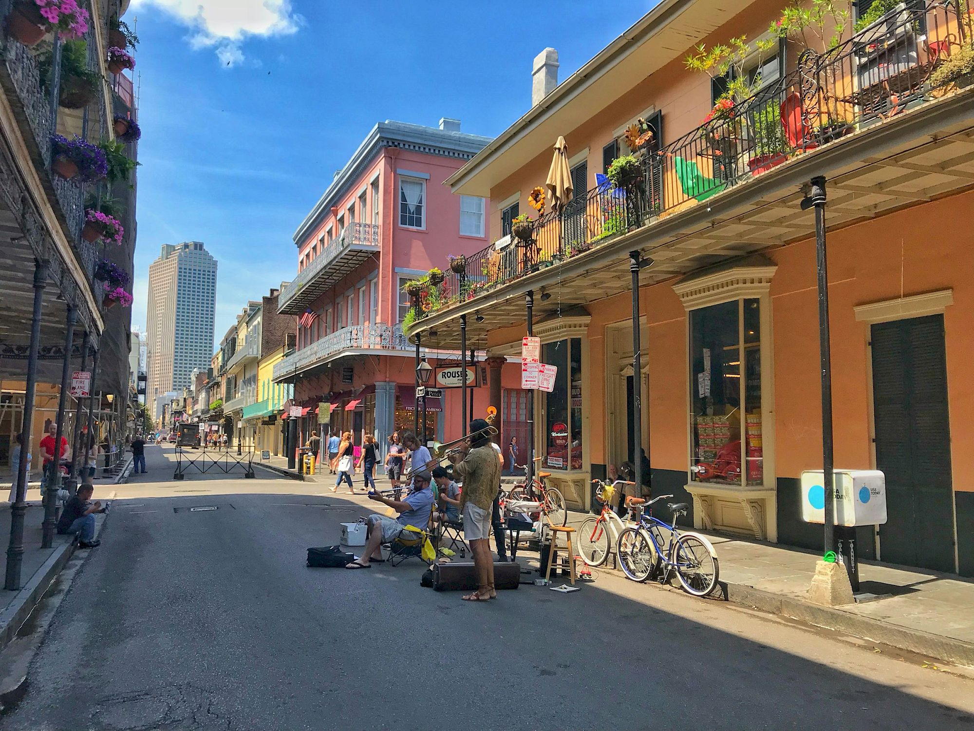 A lively street scene with musicians performing, people walking, and colorful buildings adorned with balconies on a sunny day.