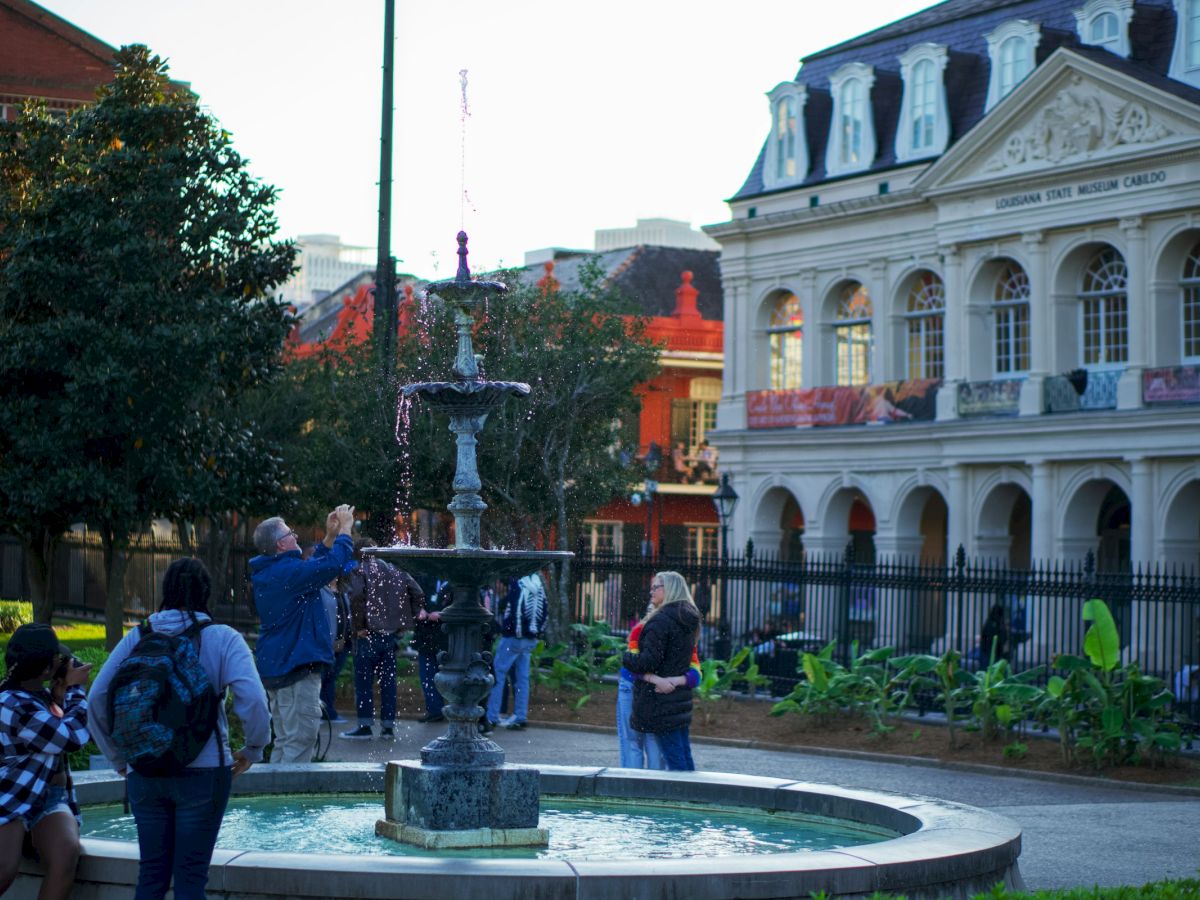 People are gathered around a fountain in a garden, with a historic building in the background, enjoying a pleasant day.