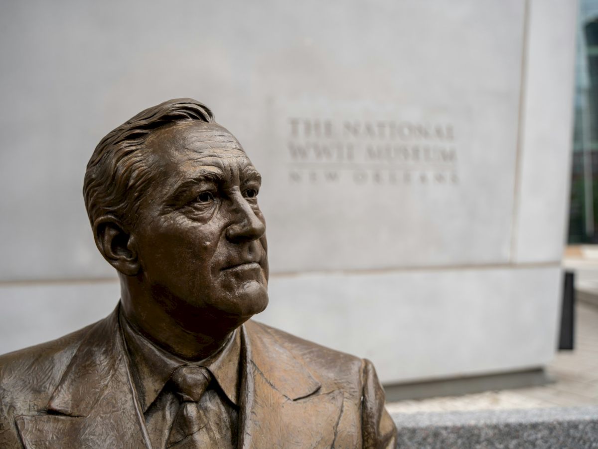 A bronze statue of a man in a suit stands outside, with "The National WWII Museum, New Orleans" engraved on the wall behind.