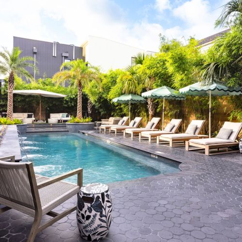 A serene poolside scene with lounge chairs, umbrellas, palm trees, and a modern pool surrounded by lush greenery and a stone-tiled deck.