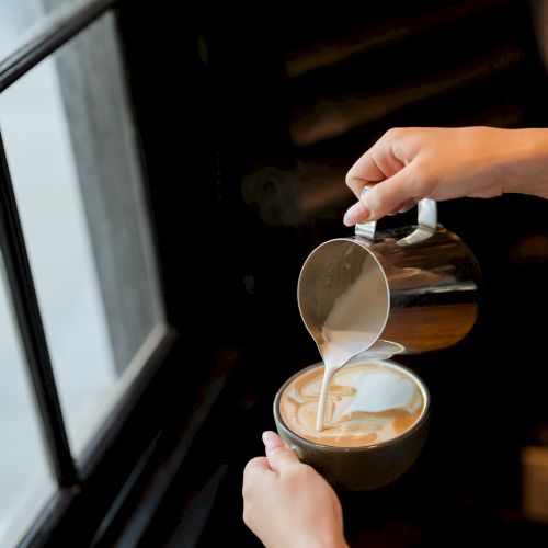 A person is pouring milk into a cup of coffee, creating latte art near a window.