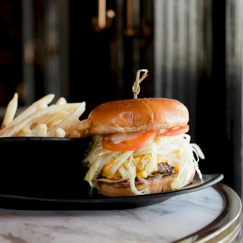 A burger with lettuce and tomato, served with fries on a dark plate, sits on a marble table.