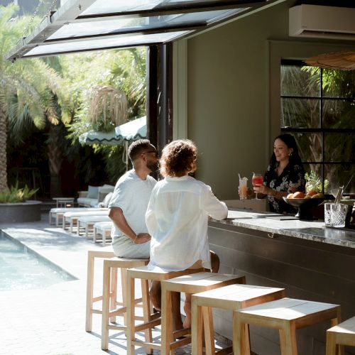A poolside bar scene with people seated on stools, enjoying drinks under an open, shaded area amidst greenery.