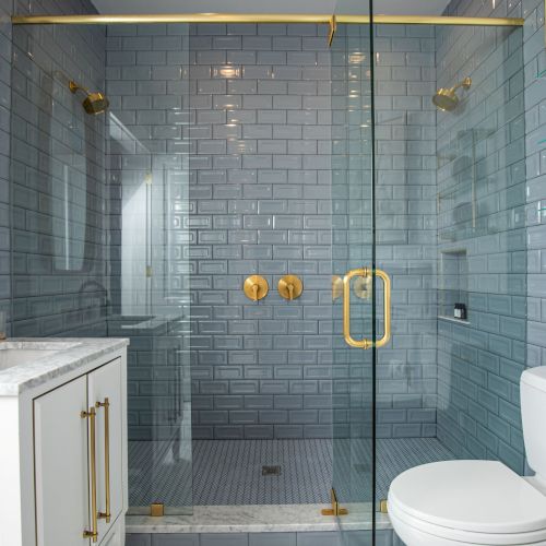 A bathroom with a glass shower, marble-topped vanity, gold fixtures, and a white toilet. Towels are neatly stacked on a wall shelf.
