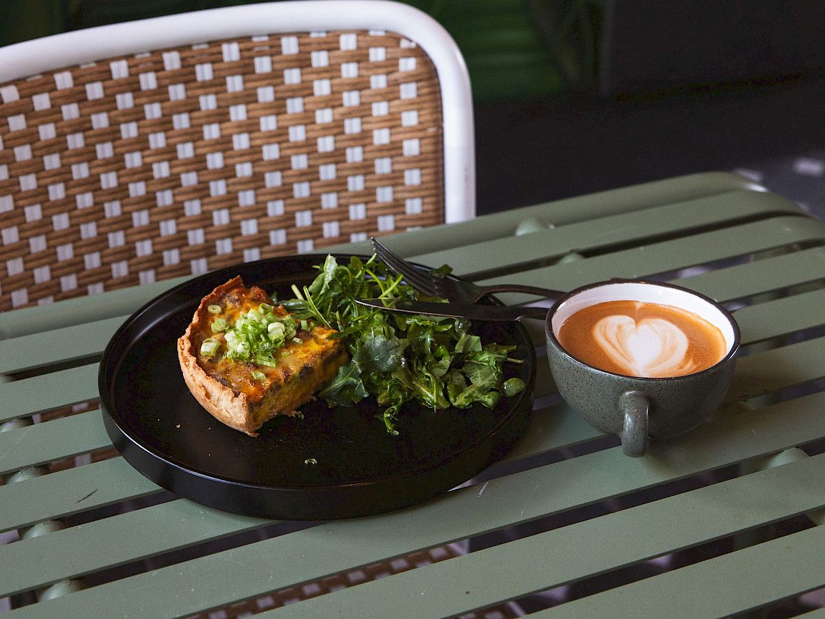 Outdoor patio brunch: quiche with greens and a latte cup, with purple flowers and green leaves in the background.