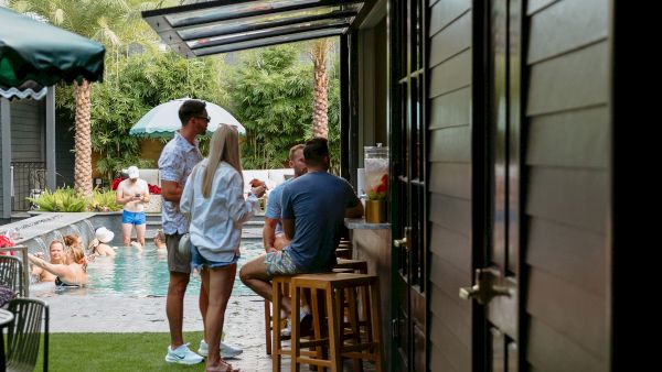 People socialize by a pool with an outdoor bar area on a paved path. The scene is relaxed with a casual outdoor setting.