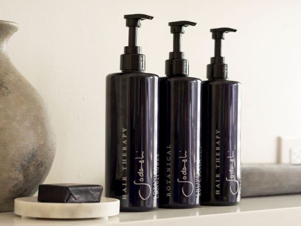 Three black pump bottles labeled "Botanical Hair Therapy" and a bar of soap on a shelf, next to a ceramic vase.