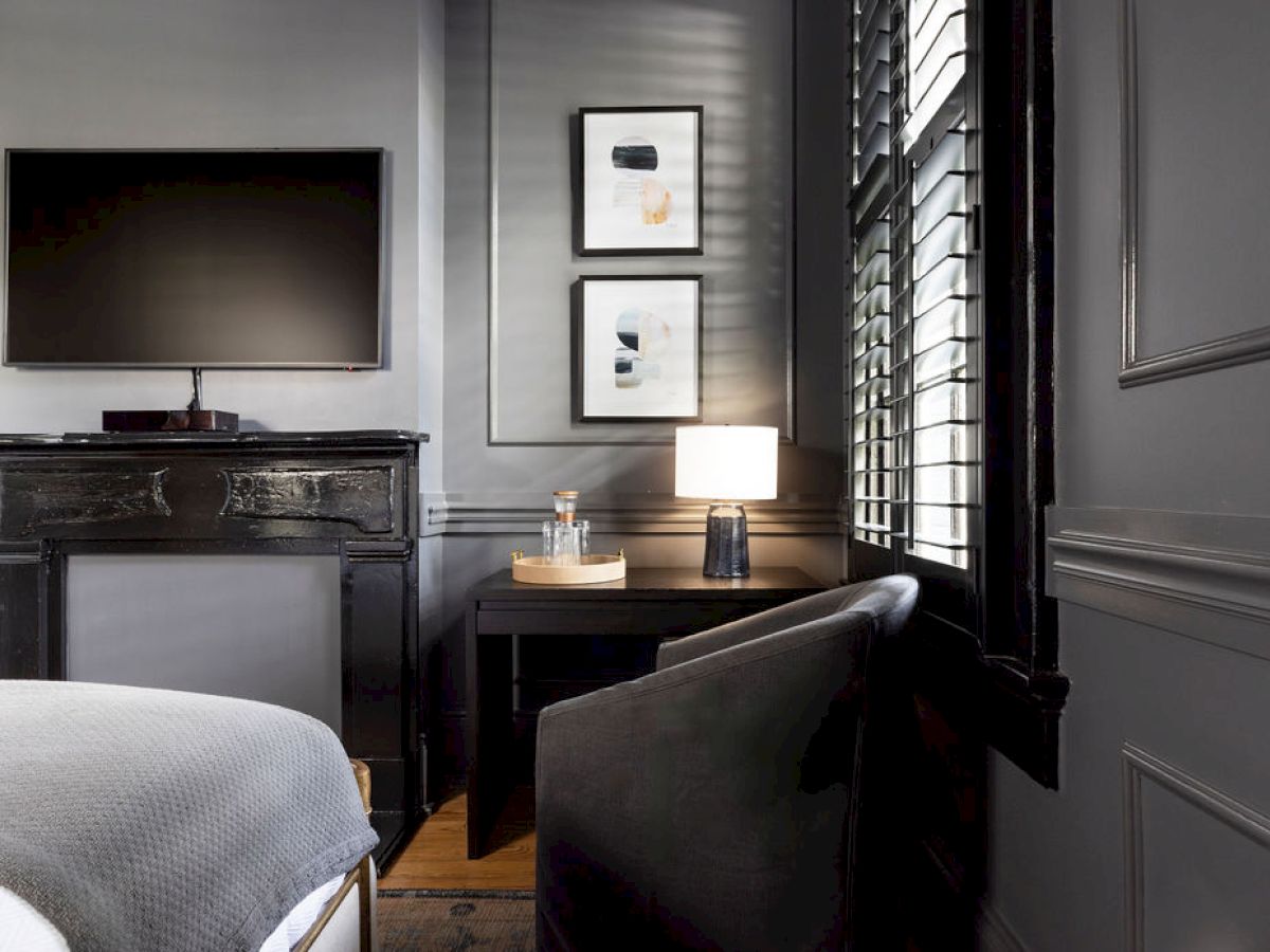 A modern room features a TV above a fireplace, two framed artworks, a desk with a lamp, and a chair beside a window with shutters.