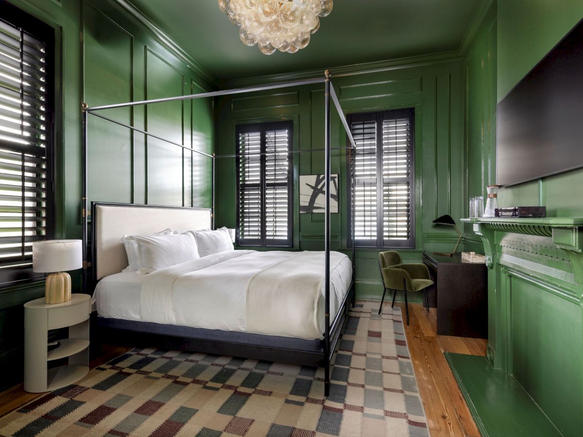 A green bedroom with a canopy bed, white bedding, patterned rug, chandelier, and a flat-screen TV; windows have shutters.