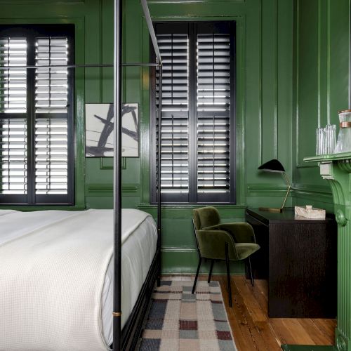 A green-walled bedroom with a canopy bed, desk, chair, and windows with shutters, creating a cozy and stylish atmosphere.