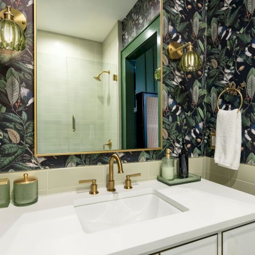 The image shows a bathroom with a floral wallpaper, white sink, gold fixtures, and green decorative elements, creating a stylish look.