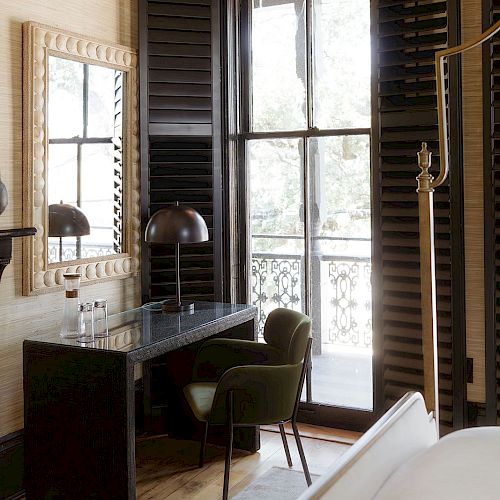 The image shows a cozy room with a desk, chair, lamp, mirror, and a large window with shutters, leading to a balcony.