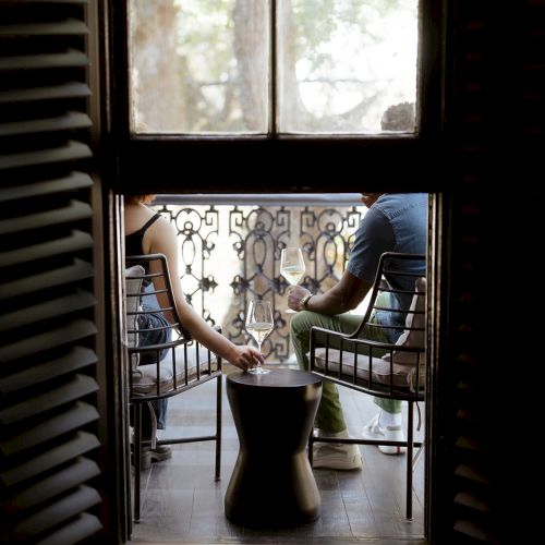 Two people sit on a balcony, visible through an open window, with a small table between them.