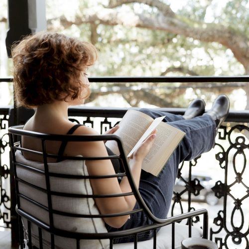 A person is sitting on a chair, reading a book with legs up on a railing, a cup on a table beside them, outdoors.