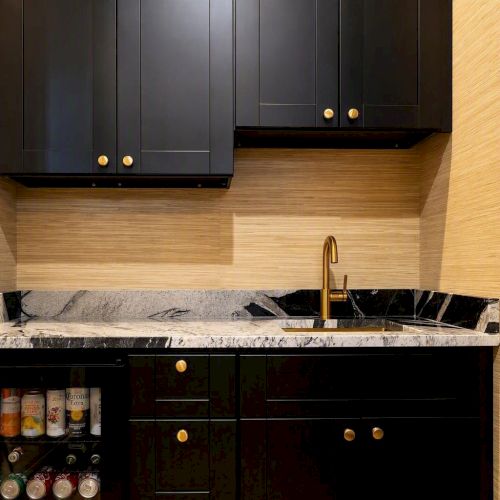 A modern kitchen sink and black cabinetry with a gold faucet, plus a small fridge stocked with drinks and cans below.