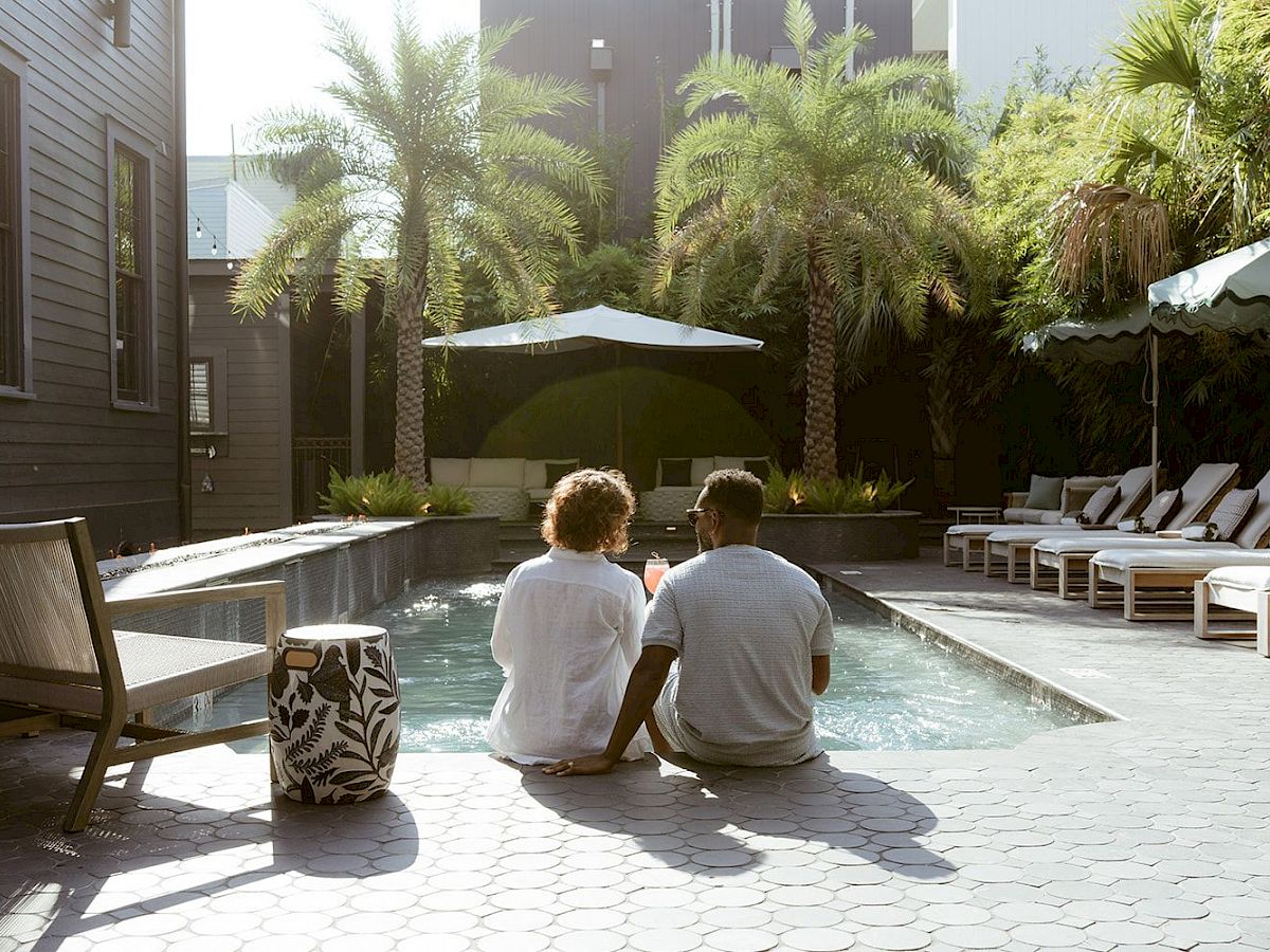 A couple sits by a pool in a sunlit courtyard, surrounded by palm trees and lounge chairs, with umbrellas providing shade.