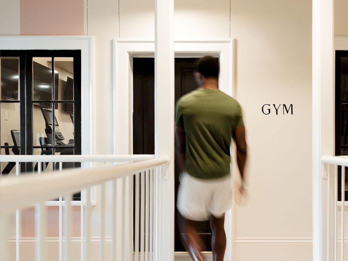 A person in athletic attire is entering a room labeled "GYM" with exercise equipment visible through windows in the background.