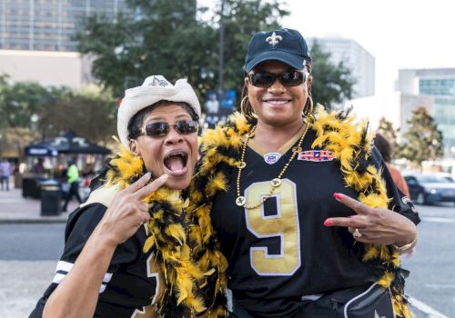 Two people wearing sports jerseys and feather boas pose energetically outdoors, with buildings and trees in the background.