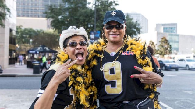 Two people wearing sports jerseys and feather boas pose energetically outdoors, with buildings and trees in the background.