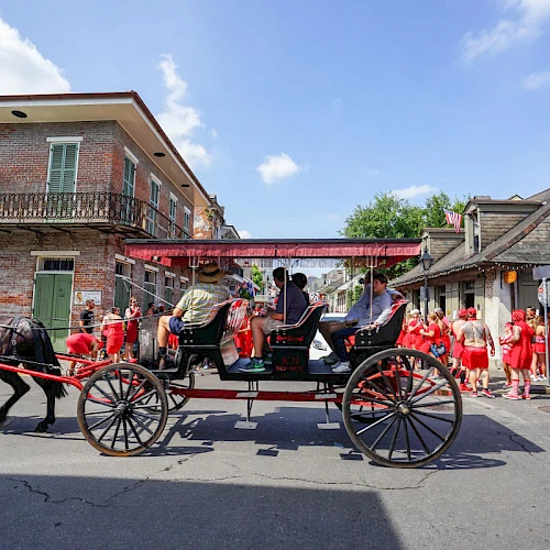 A horse-drawn carriage travels down a street with people dressed in red walking nearby, under a clear blue sky.