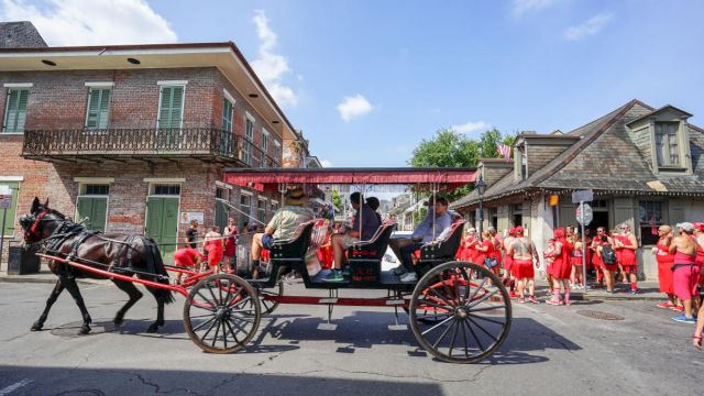 A horse-drawn carriage travels down a street with people dressed in red walking nearby, under a clear blue sky.
