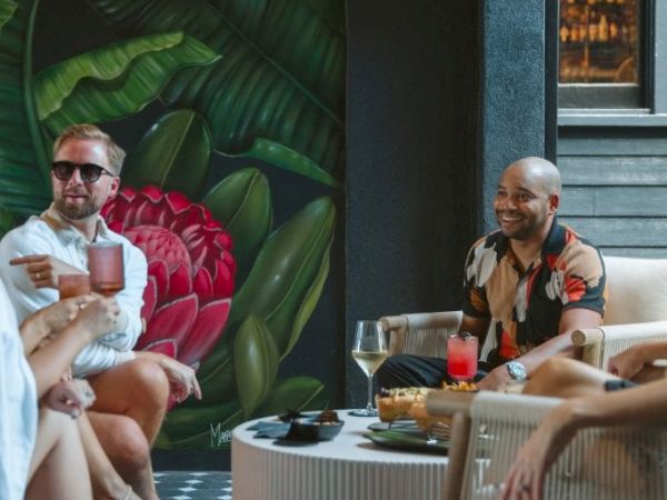 A group of people are enjoying drinks and conversation outdoors, with a large mural of leaves and a flower in the background.