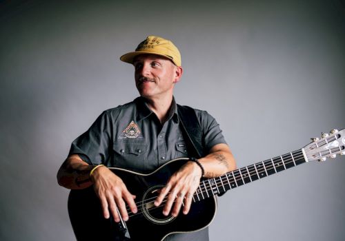 A person in a cap and casual shirt holds an acoustic guitar, looking off to the side in a neutral, softly lit setting.