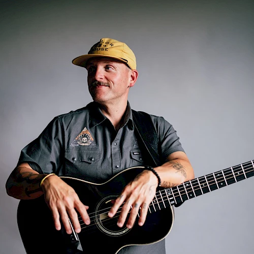 A person in a cap and casual shirt holds an acoustic guitar, looking off to the side in a neutral, softly lit setting.