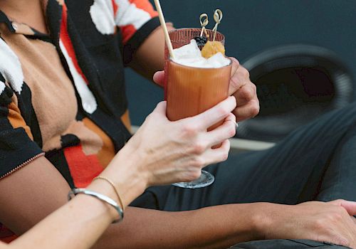 Two people are holding a cocktail garnished with a slice of fruit and skewers, one wearing a colorful shirt.