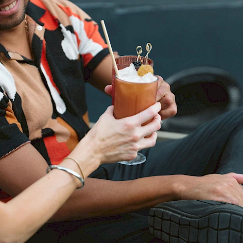 Two people are holding a cocktail garnished with a slice of fruit and skewers, one wearing a colorful shirt.