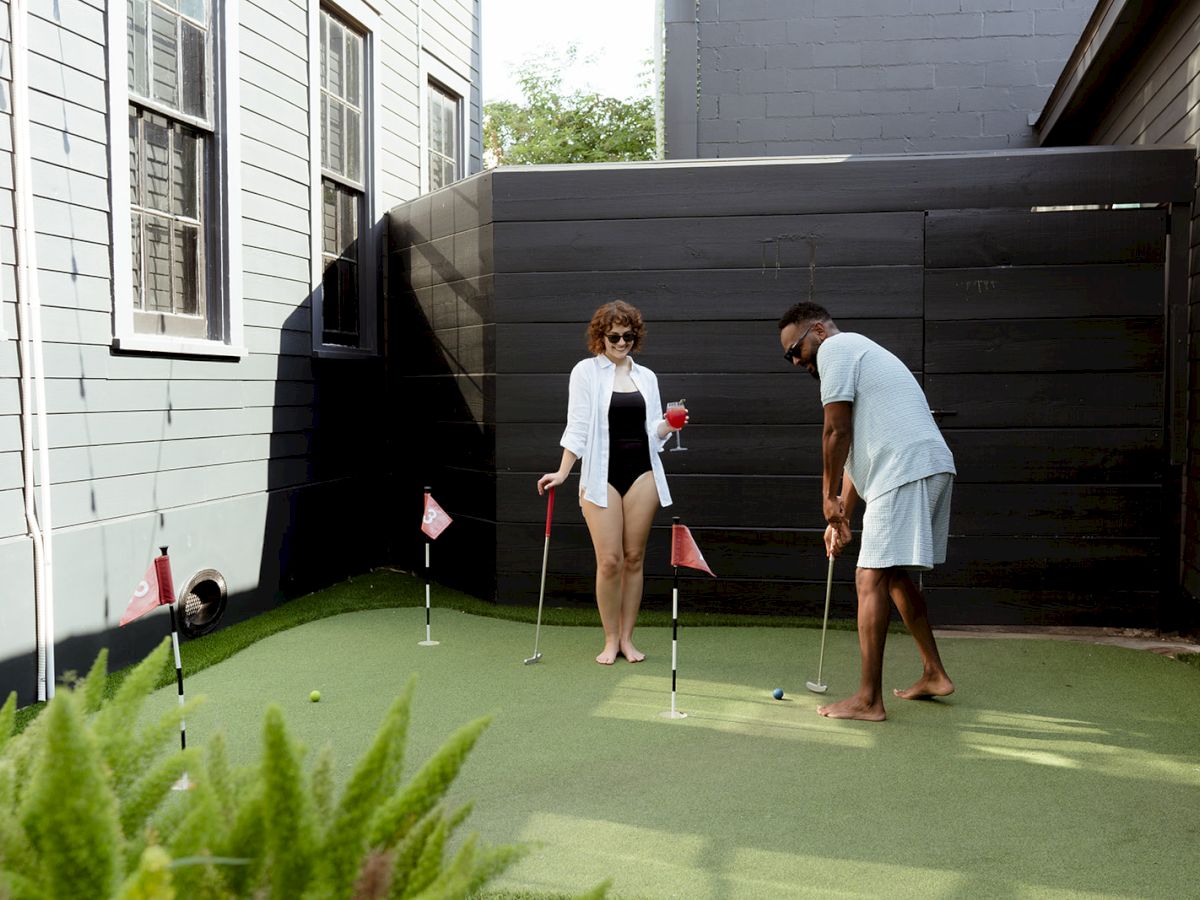 Two people are playing mini-golf on an outdoor putting green, surrounded by buildings.