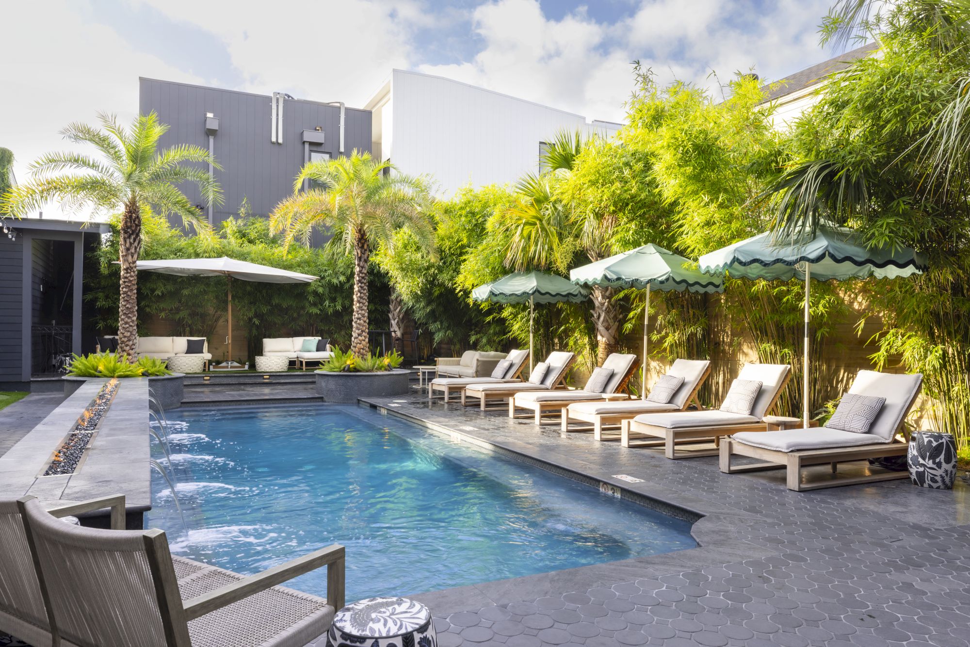 A serene outdoor pool area with lounge chairs, umbrellas, palm trees, and lush greenery under a blue sky.