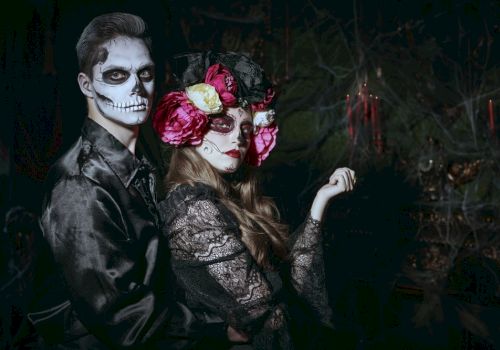 A couple in dark clothing with skull makeup. The woman wears a flower crown. The background features dark, eerie lighting and candles.