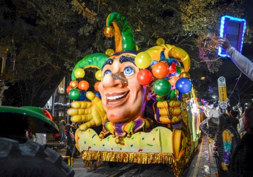 A colorful parade float with a jester face and balloons is being photographed by a crowd under night lights.