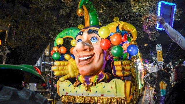 A colorful parade float with a jester face and balloons is being photographed by a crowd under night lights.