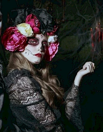 A person with intricate face paint and floral headpiece in a dark, decorative setting.