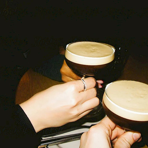 Two people clinking glasses filled with a creamy, frothy drink in a dimly lit setting.